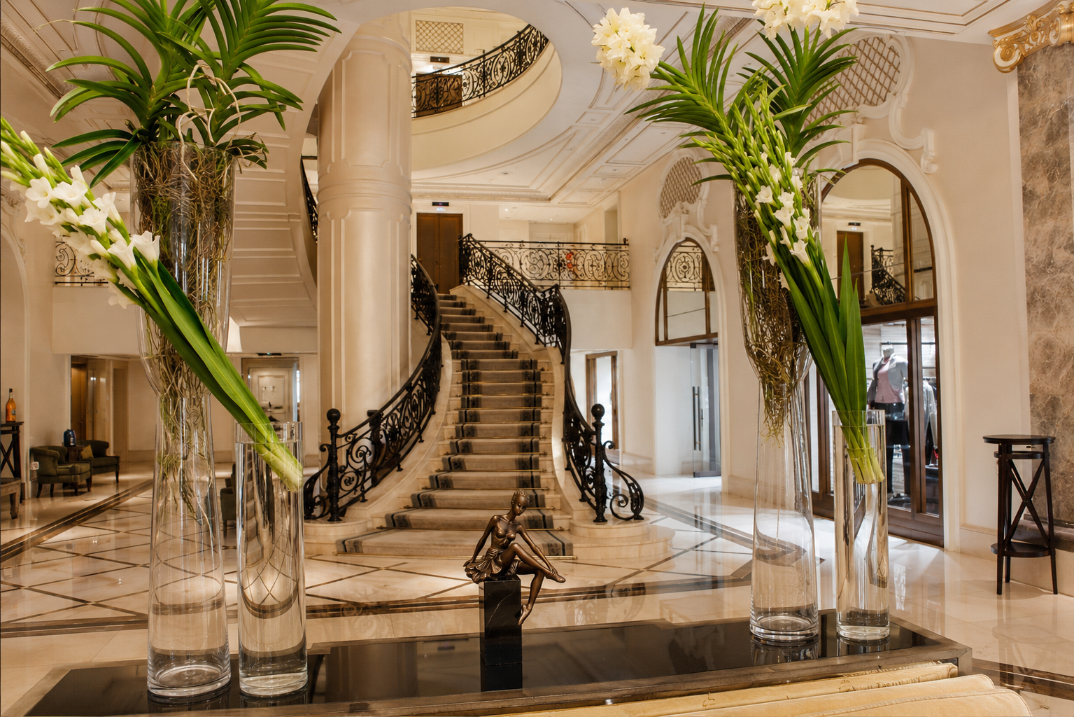 A grand hotel lobby with a sweeping staircase, marble floor and tall floral arrangements
