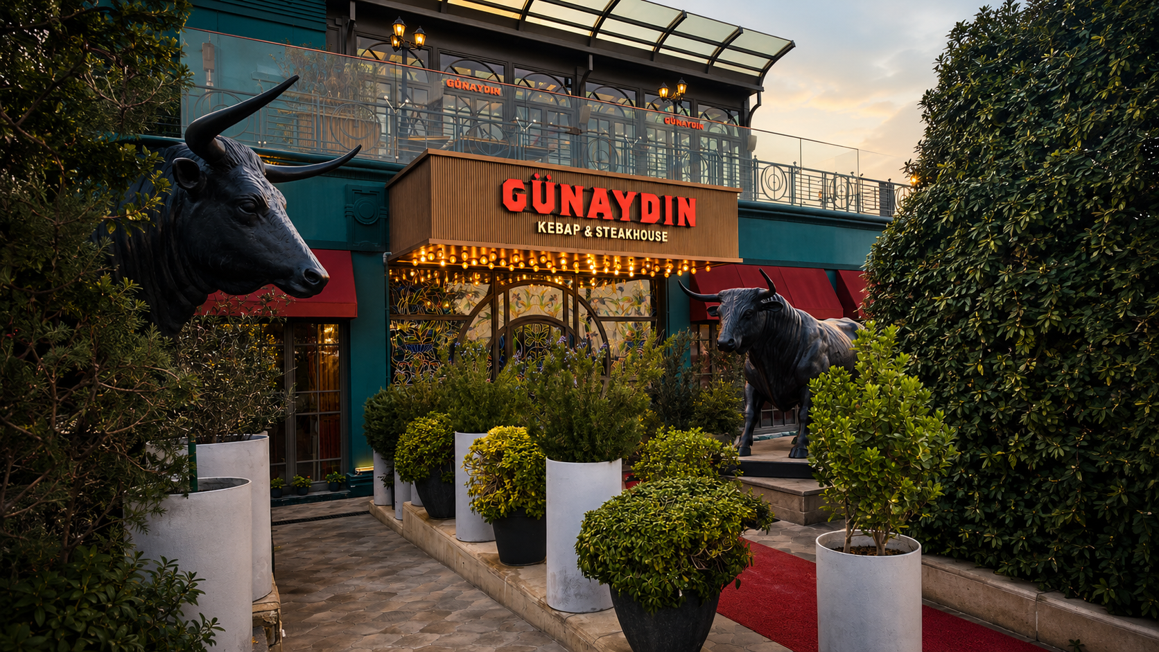 The entrance of Günaydın Kebap & Steakhouse in Baku at dusk, framed by topiary and two black bull sculptures